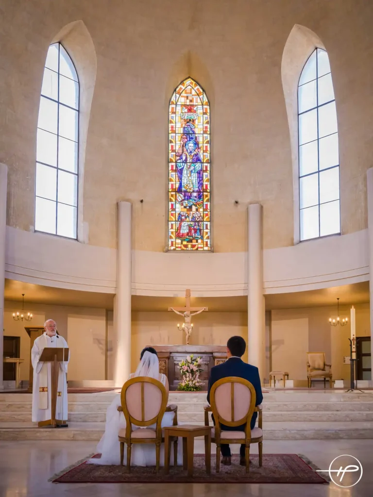 Vaste église, autel et vitrail mis en valeur. Avec les mariés assis au pied de l’autel pour la cérémonie religieuse de leur union célébrée par le prêtre debout devant un pupitre et faisant face aux futurs mariés.