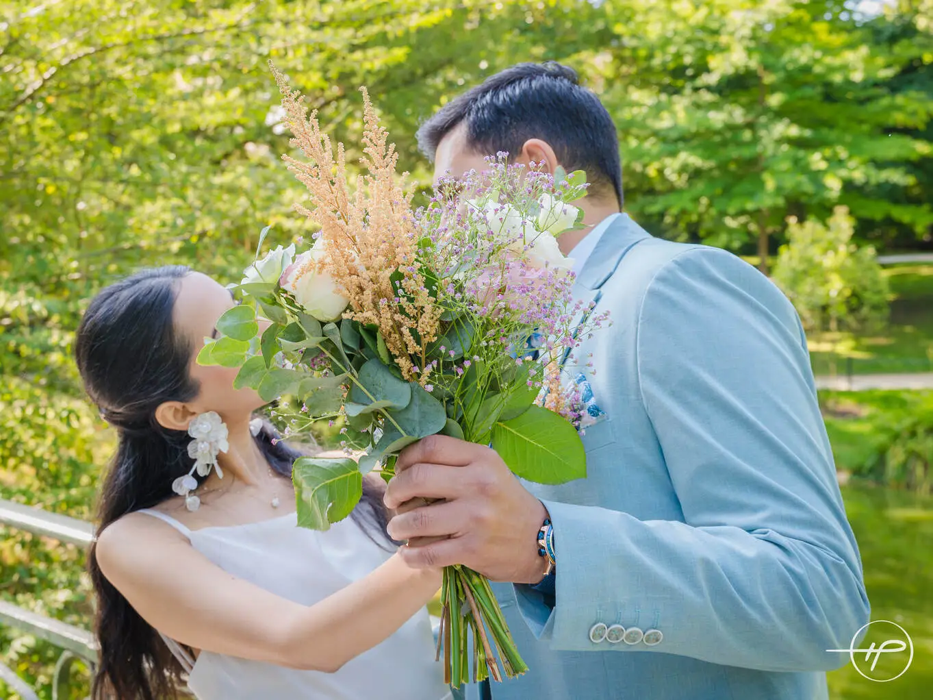 Couple de mariés s’embrassant dans un parc verdoyant à Paris, cachés derrière un bouquet de mariage romantique aux fleurs blanches, roses et feuillages.
