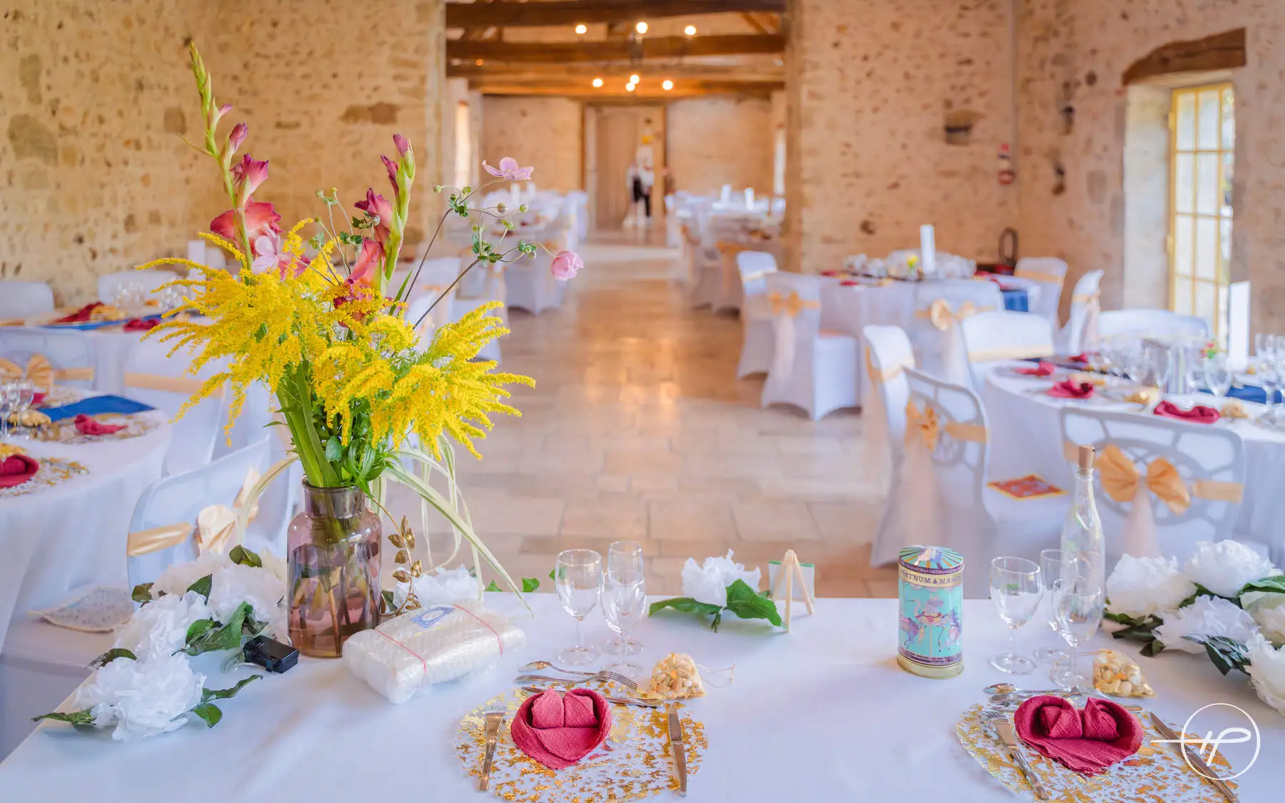 Salle de réception de mariage décorée avec élégance : tables rondes nappées de blanc, chaises avec nœuds dorés, serviettes pliées en cœur et centre de table floral coloré, idéale pour un mariage romantique.